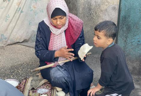 صورة لأمرأة فلسطينية في أحدمراكز الإيواء تحاول إعداد الخبز لإطعام طفلها في ظل انتشار المجاعة بسبب التصعيد العسكري ضد قطاع غزة – قطاع غزة لعام 2024