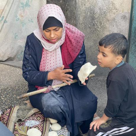 صورة لأمرأة فلسطينية في أحدمراكز الإيواء تحاول إعداد الخبز لإطعام طفلها في ظل انتشار المجاعة بسبب التصعيد العسكري ضد قطاع غزة – قطاع غزة لعام 2024